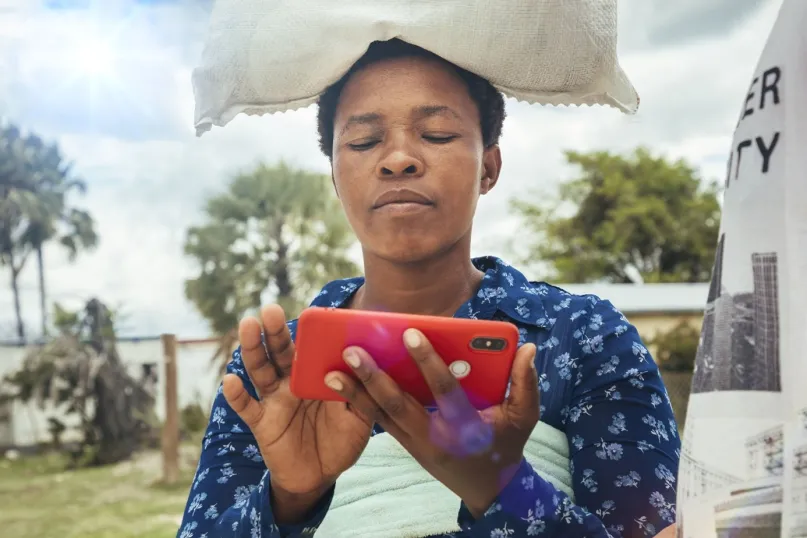 Woman with a sack balanced on her head touching a mobile phone.