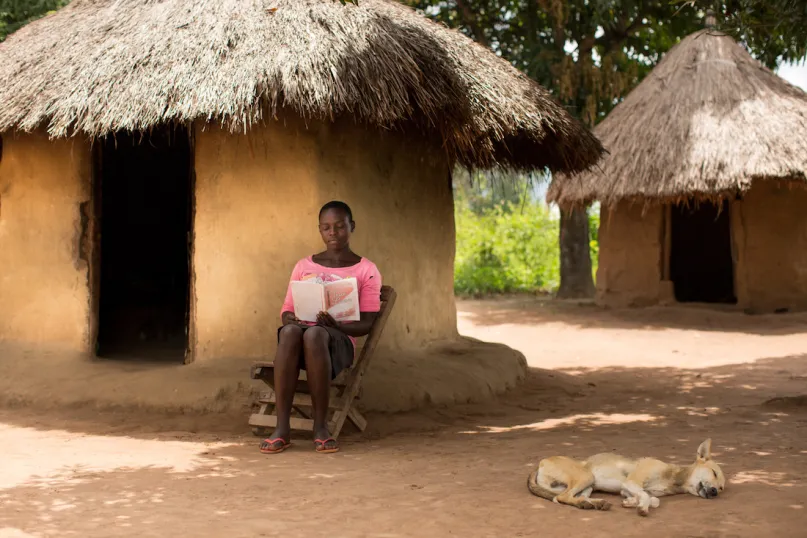 A woman reads her book while her dog sleeps next to her at a family compound in Uganda.