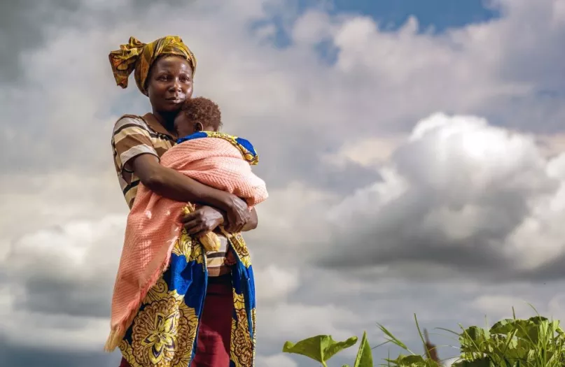 African woman holding baby in field with clouds behind her.