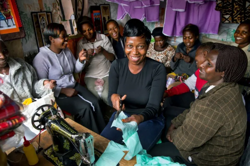 Group of women from Kenya smiling, sewing and cutting cloth by a sewing machine.