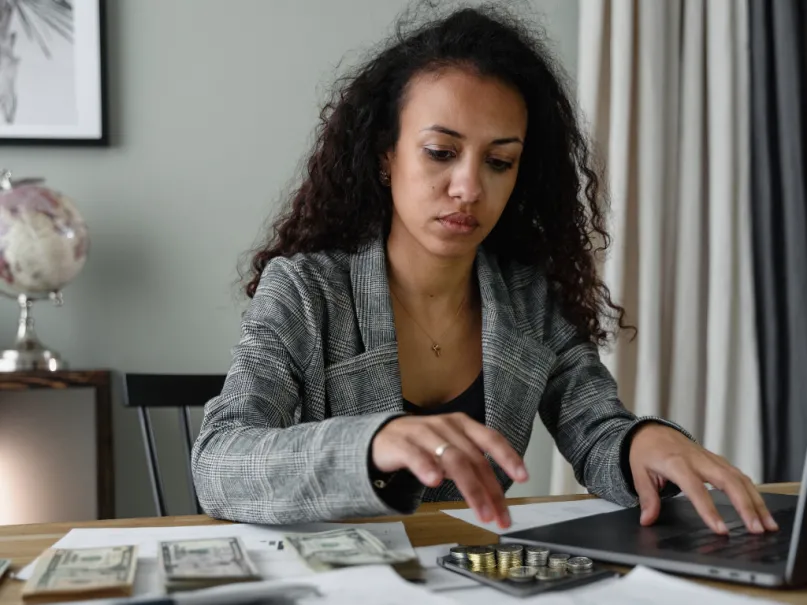 إمرأة في سترة منقوشة تجلس أمام حاسوبها وتجري حسابات مالية وعلى الطاولة أمامها كدسات من النقد الورقية والمعدنية.