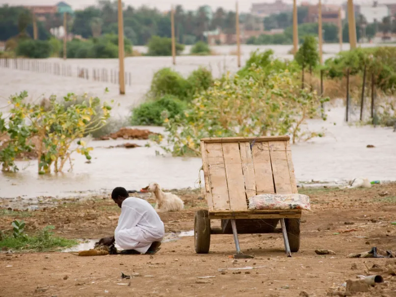 رجل بلباس أبيض يعمل في أرض فيها نبات متناثر في السودان وإلى جانبه عجلة يجر عليها أغراضه.