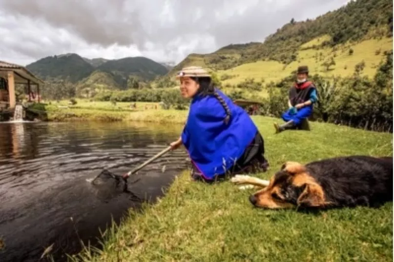 Una mujer arrodillada junto a un río, pescando, junto a ella está un hombre, y un perro acostado, en Colombia.