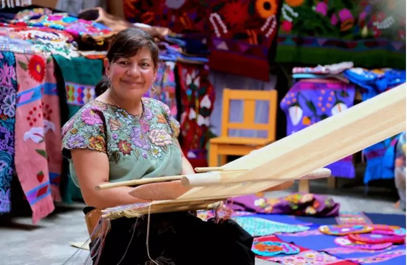 Mujer sentada tejiendo en un telar entre artesanías textiles en México.
