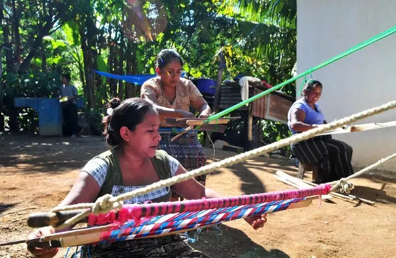 Tres mujeres sentadas tejiendo en telares artesanales en Guatemala.