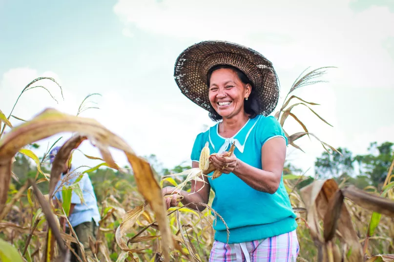A woman smiles in a field holding a piece of corn