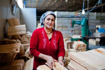 إمرأة لابسة كنزة حمراء وحطة على رأسها تعمل في مصنع للأخشاب أمامها طاولة عليها لوحات خشبية مستطيلة.
