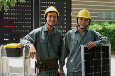Two young men with yellow helmets smiling and holding solar panels.