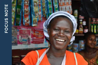 Smiling woman with apron and head covering in front of a small store.