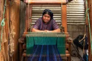Guatemalan woman leaning on a loom with green and blue textiles.