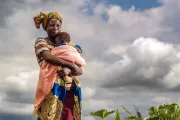 African woman holding baby in field with clouds behind her.