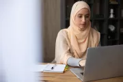 امرأة مسلمة بالحجاب تركز على عملها جالسة على طاولة مع جهاز كمبيوتر محمول ودفتر منظم أمامها.